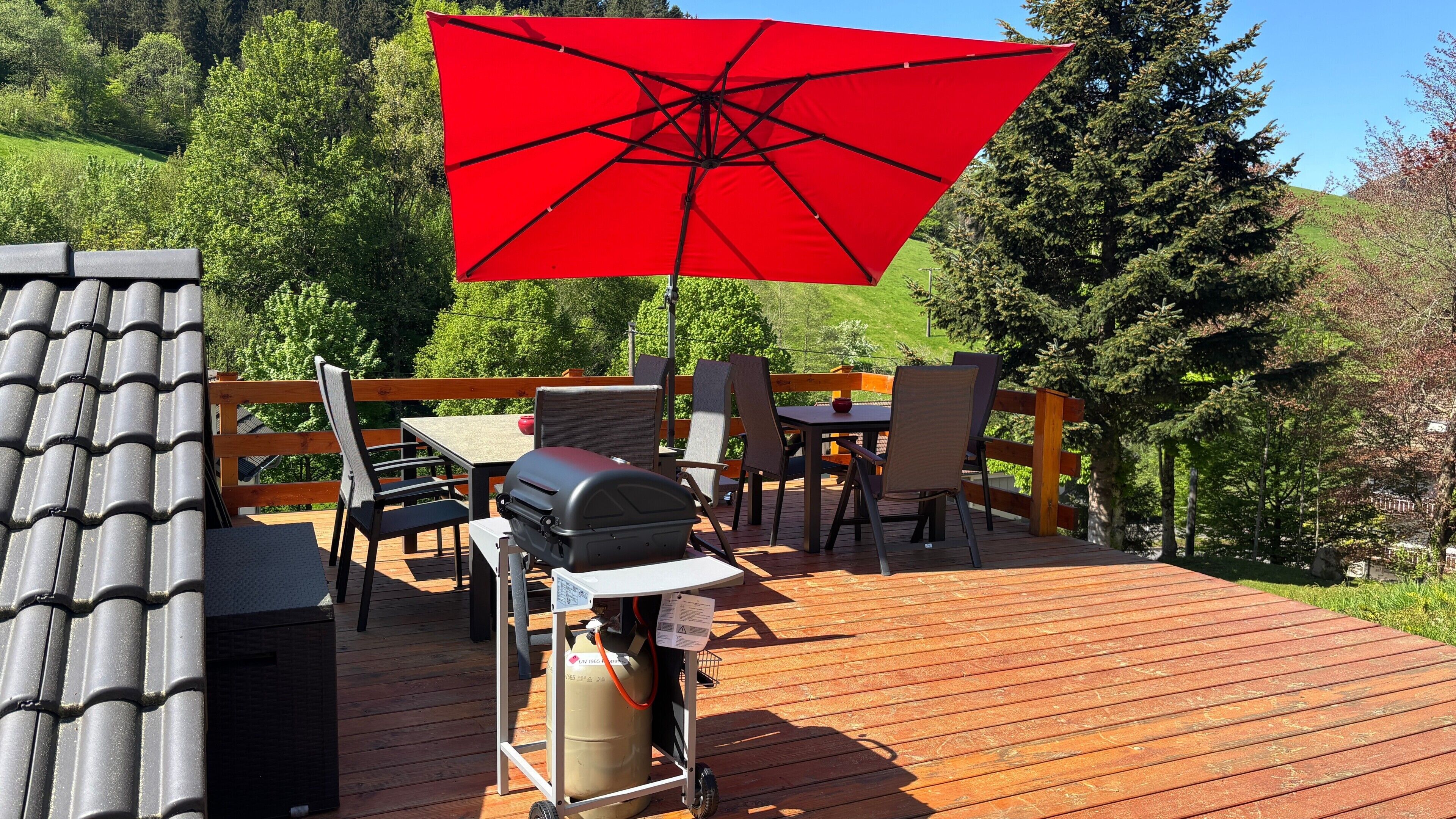 Eine Holzterrasse mit Gartenmöbeln, einem Grill und einem roten Sonnenschirm.