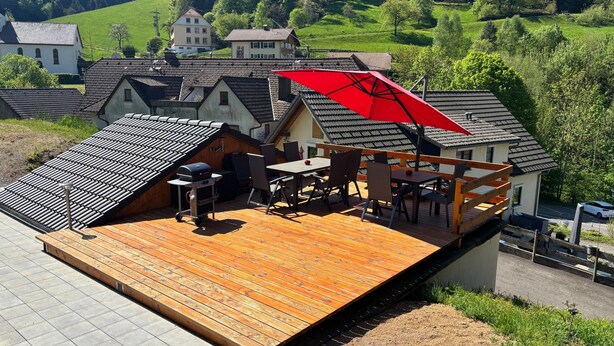 A wooden deck with a barbecue grill and outdoor seating, overlooking a residential area with houses and greenery.