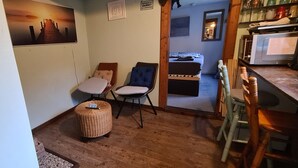 A cozy room with a wooden floor, two chairs with cushions, a small ottoman, a bed with a wooden headboard, and a kitchen area with a microwave. - Tiny house in the Wingster Forest, near Nordesse, peace & relaxation in nature (Wingst)