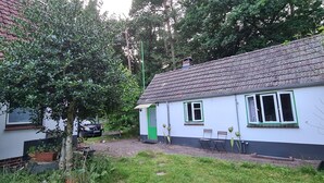 A small house with a tiled roof, a green door, and a window with a white frame. There is a chair and a table outside, and a tree is visible in the foreground. - Tiny house in the Wingster Forest, near Nordesse, peace & relaxation in nature (Wingst)