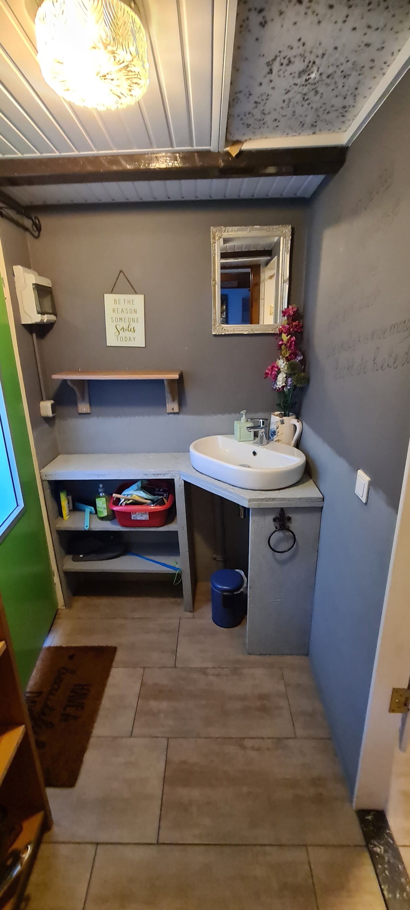 A compact bathroom with a white sink, a mirror, a shelf with toiletries, and a small trash bin.
