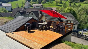 Une terrasse en bois équipée d’un barbecue et de meubles d’extérieur, offrant une vue sur un quartier résidentiel avec ses maisons et sa verdure.