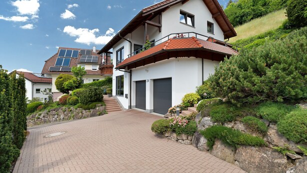 A modern two-story house with a paved driveway, a garage, and a balcony with plants.