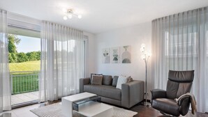 Ein modernes Wohnzimmer mit einer Couch, einem Relaxsessel, einem Couchtisch und Blick auf eine grüne Landschaft durch große Fenster.
