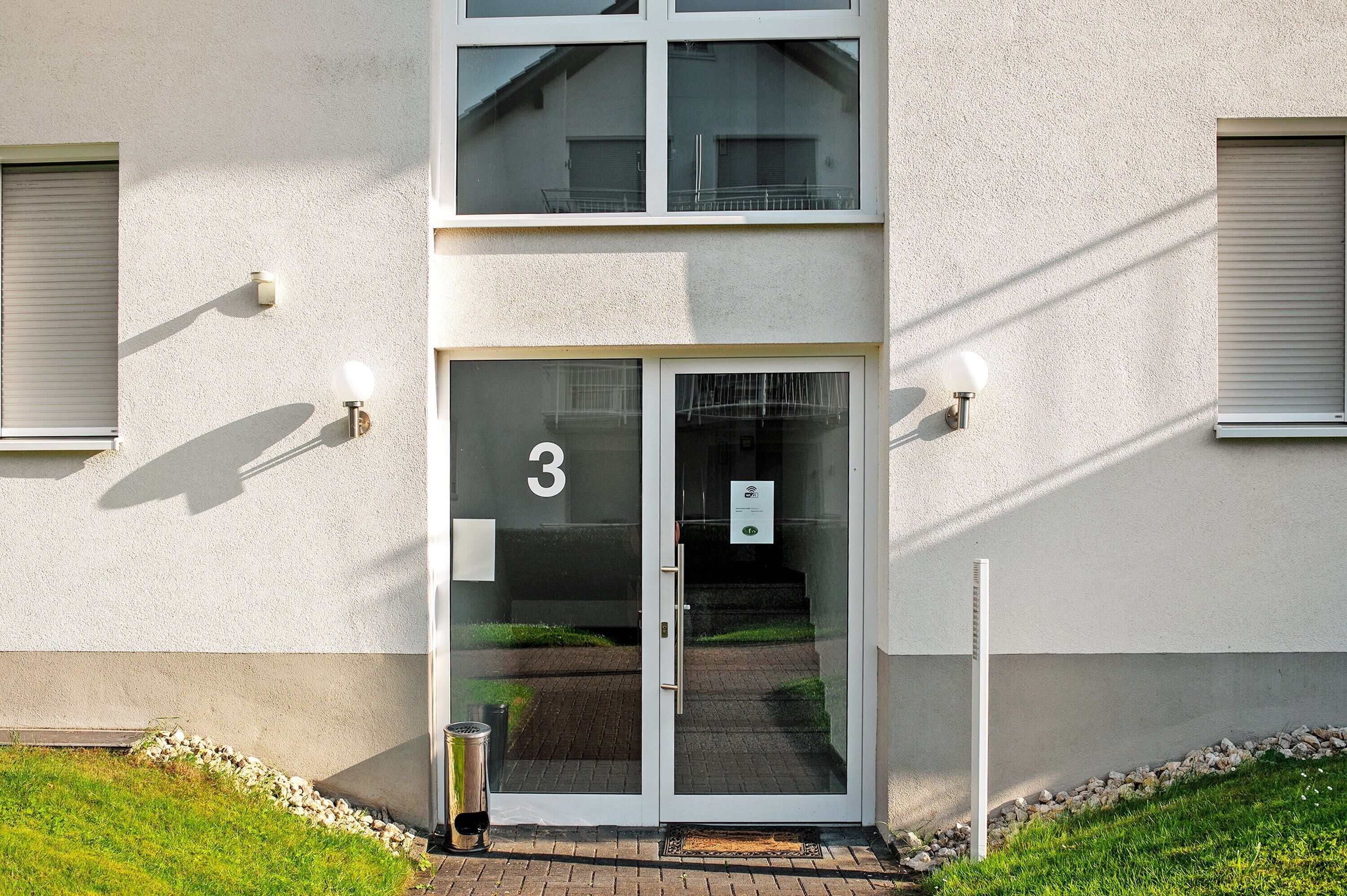 Ein Gebäudeeingang mit Glastür, der Hausnummer '3' auf der Tür und einem kleinen Schild an der Wand.