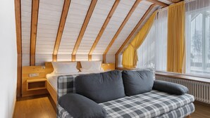 Una habitación con techo inclinado, una cama con ropa de cama gris, un sofá escocés, una mesita de noche de madera y una ventana con cortinas amarillas.