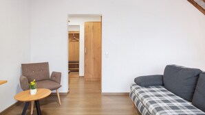 Habitación con sofá, una silla, una mesita con un florero, suelo de madera y estructura de techo de madera.