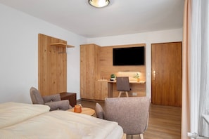 A modern hotel room with a bed, two armchairs, a small table, a TV, a wooden wardrobe, and a kitchenette.
