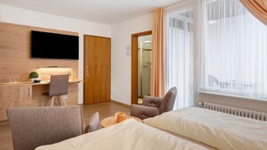 Habitación de hotel con cama, dos sillones, escritorio con televisor y zona de ducha.