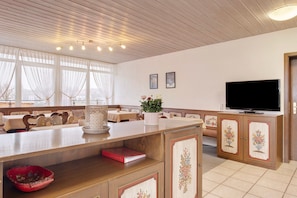 A hotel room with a wooden ceiling, a television, a dining area with tables and chairs, and a large window with curtains.