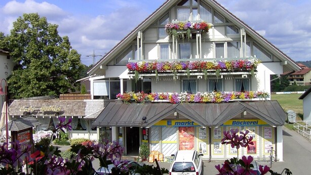 A two-story building with a gabled roof, storefronts labeled 'E. Markt' and 'Bäckerei,' and flower boxes on the balconies.