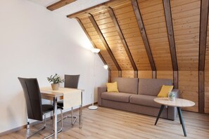 A loft-style room with a sloped ceiling, a dining table with chairs, a sofa with cushions, and a small round table with a lamp.