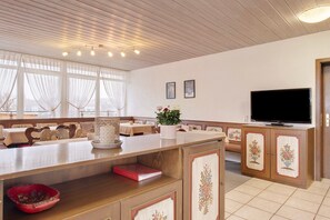 Une chambre d’hôtel avec un plafond en bois, une télévision, un coin repas avec des tables et des chaises, et une grande fenêtre avec des rideaux.