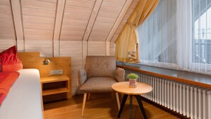A cozy room with a wooden ceiling, a bed with a red blanket, a small wooden table with a plant, and a window with curtains.