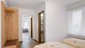 A bedroom with a wooden bench, a bed with floral bedding, a window with sheer curtains, and a view into a bathroom. - Apartment '2-zimmer- Nr 7' with Balcony and Wi-Fi (Ühlingen-Birkendorf)