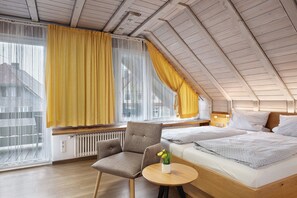 A modern loft bedroom with a sloped ceiling, a large window with yellow curtains, a wooden floor, a single bed, a chair, and a small table.