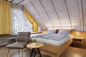 A bedroom with a sloped ceiling, a bed with two pillows, a chair, a small table with a plant, and a window with yellow curtains.