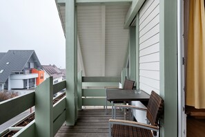 A covered outdoor seating area with a table and chairs, a wooden floor, and a view of neighboring buildings.