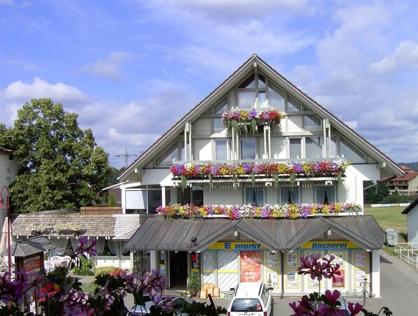 A two-story building with a gabled roof, storefronts labeled 'E. Markt' and 'Bäckerei,' and flower boxes on the balconies.
