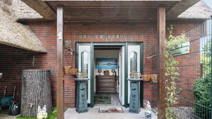 A brick building with a thatched roof entrance, a wooden bench, and a small garden area with a dog statue. - Apartment 'Lütt Stuv' with Private Terrace, Shared Garden, and Wi-Fi (Fockbek)