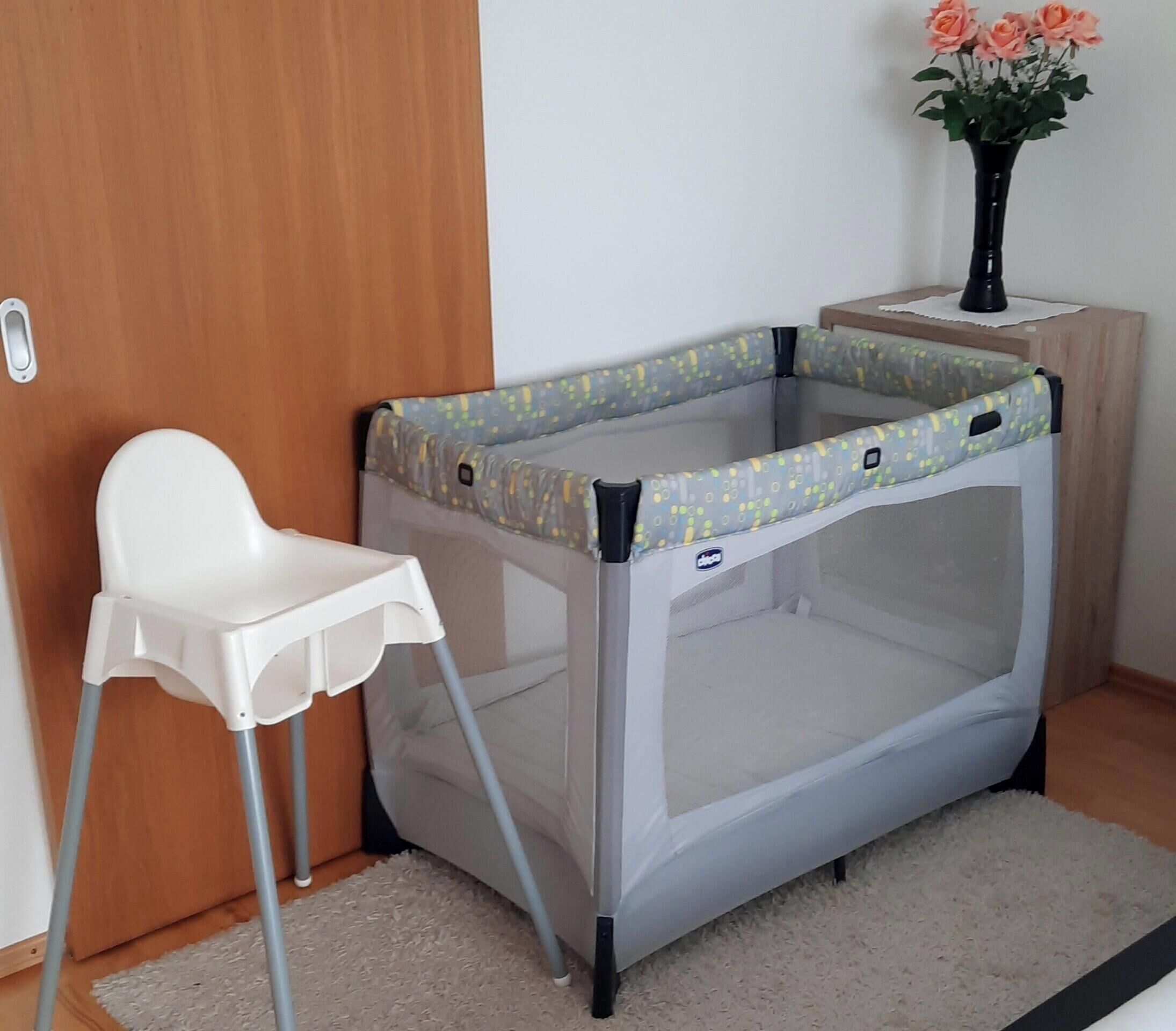 Ein Babyreisebett mit einem weißen Stuhl, einem kleinen Tisch mit Blumen in einer Vase und einer Holztür.