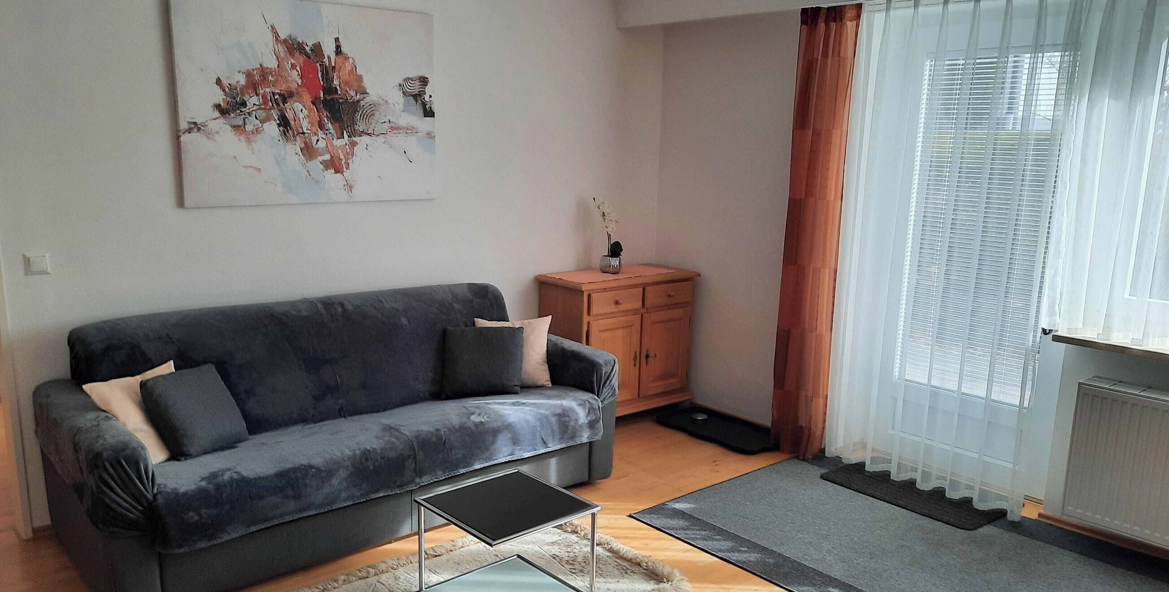 Un salon avec un canapé gris, un petit meuble en bois, une table basse en verre et un grand tableau abstrait accroché au mur.