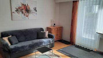 Un salon avec un canapé gris, un petit meuble en bois, une table basse en verre et un grand tableau abstrait accroché au mur.
