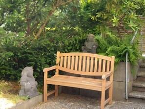 A wooden bench in a garden with dense foliage and a stone statue in the background. - Apartment 'Relax' with Mountain View, Private Garden and Wi-Fi (Bad Wildbad)
