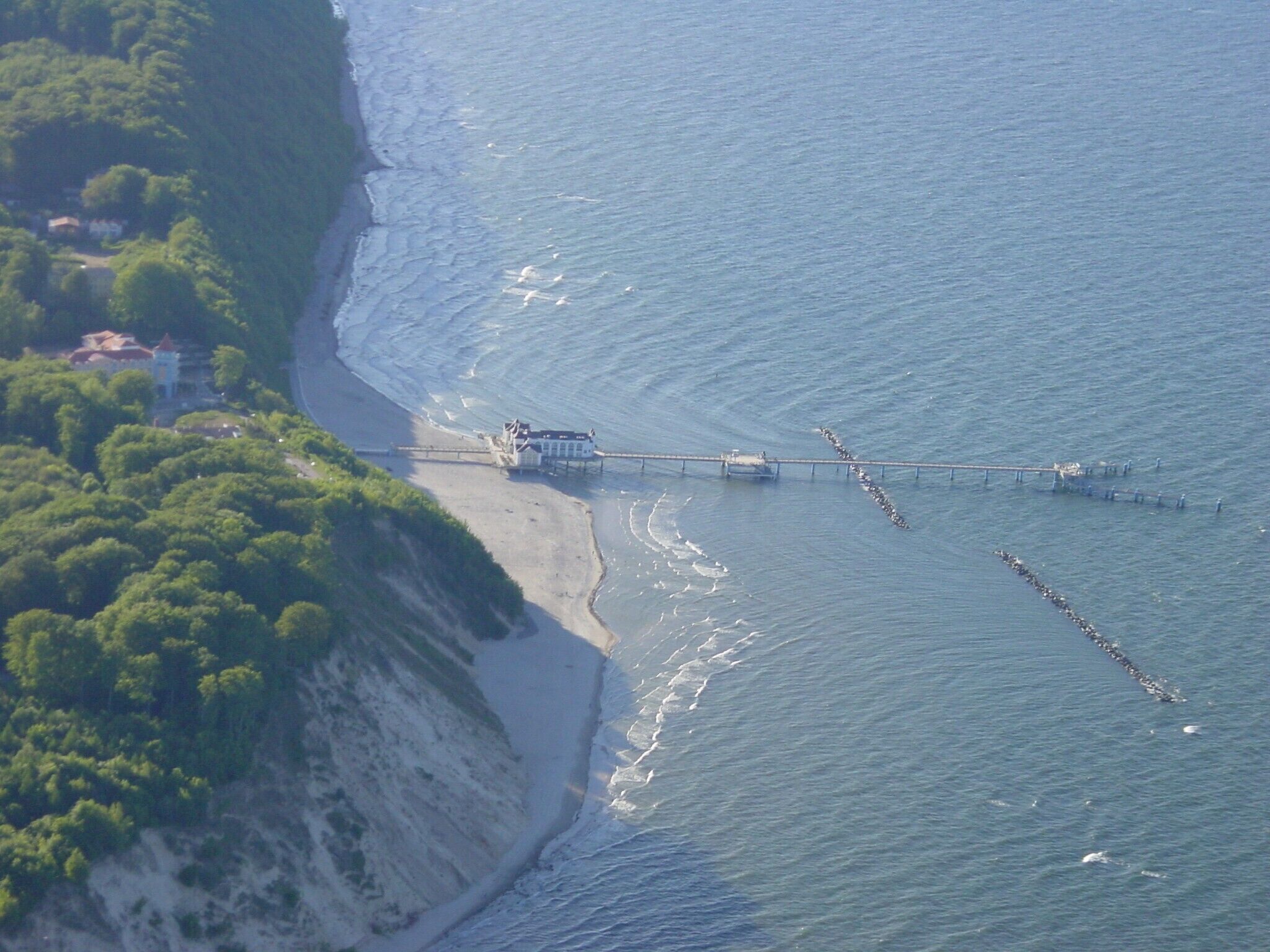 An aerial view of a coastal area with a pier extending into the sea, surrounded by greenery and a sandy beach.