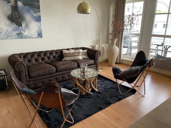 A living room with a brown sofa, two chairs, a glass coffee table, a large vase, and a painting of a lighthouse.