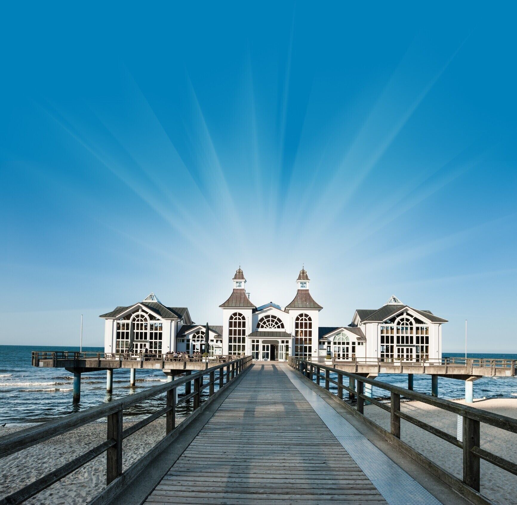 En bro, der fører hen til en række hvide bygninger med buede vinduer og et centralt tårn, med udsigt over havet.