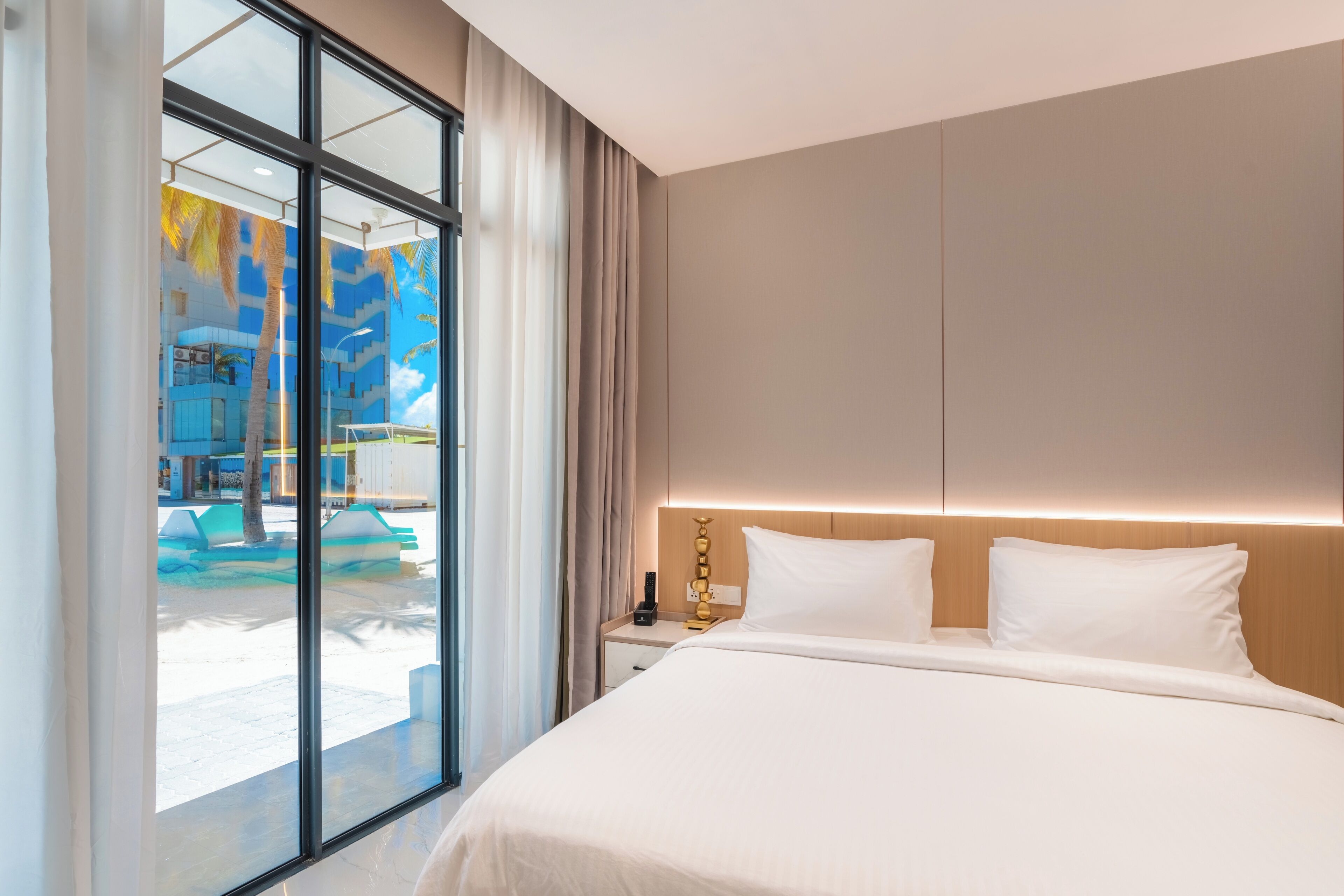 A modern hotel room with a large bed, a nightstand, and a view of a pool area through floor-to-ceiling windows.