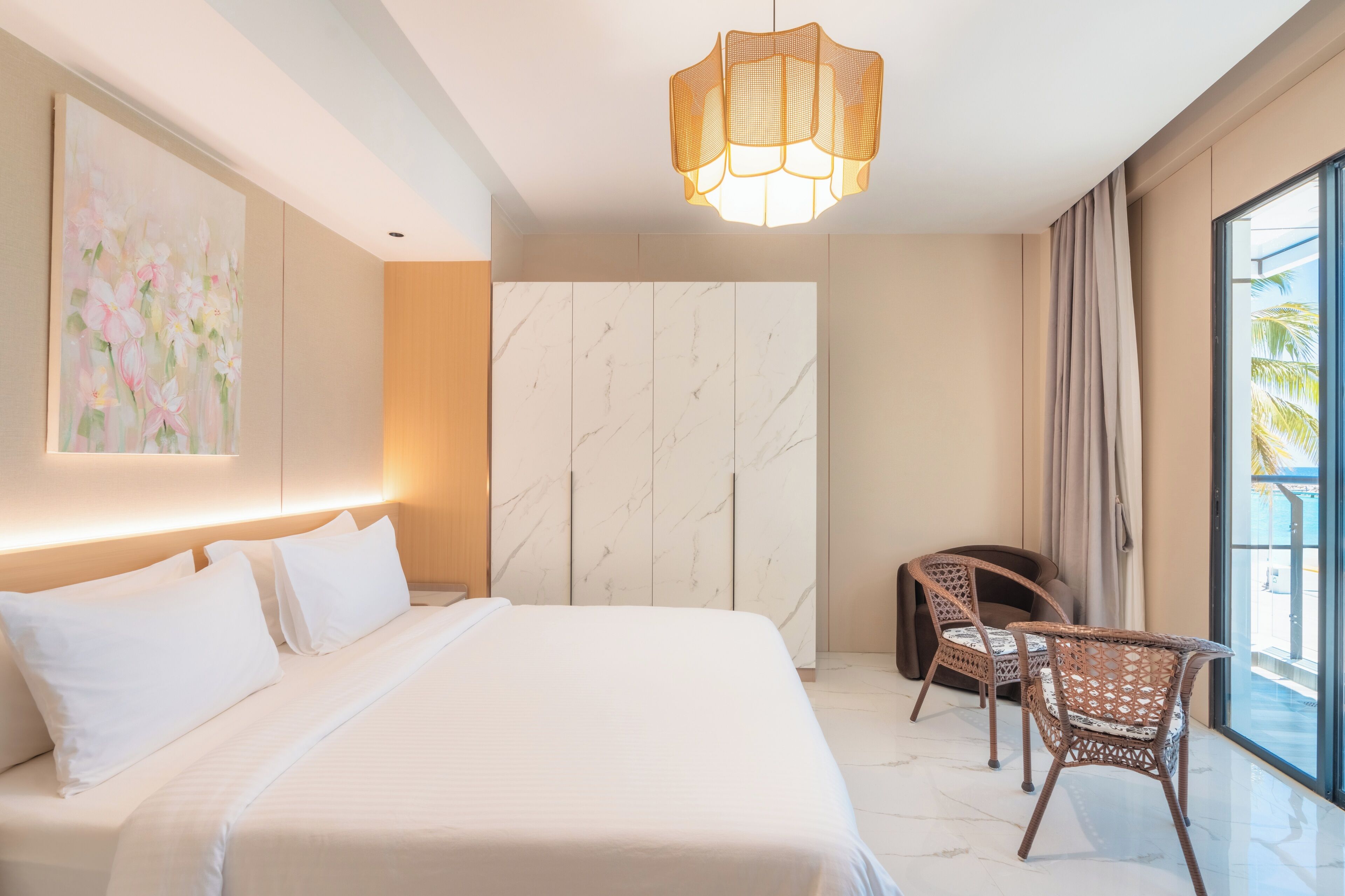 A modern hotel room with a large bed, a seating area with wicker chairs, a marble wall, and a large window with a view.