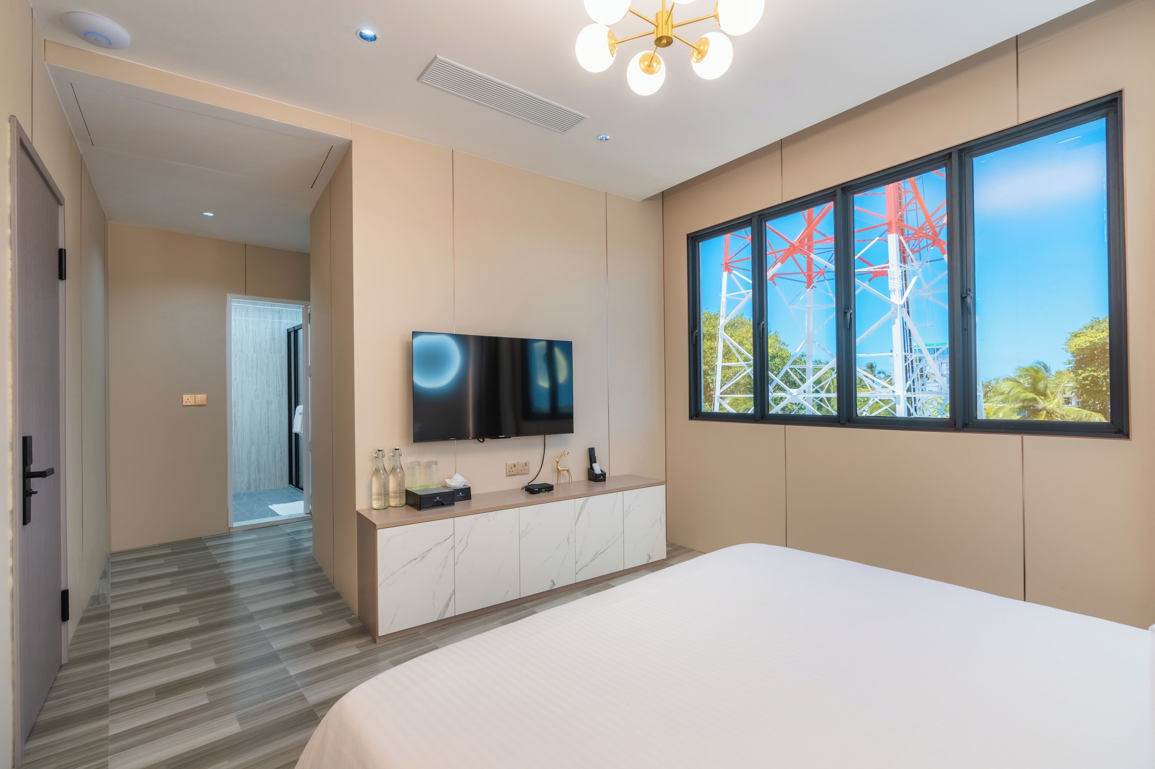 A modern hotel room with a large bed, a TV mounted on a marble cabinet, a window with a view of power lines, and a hallway leading to another room.