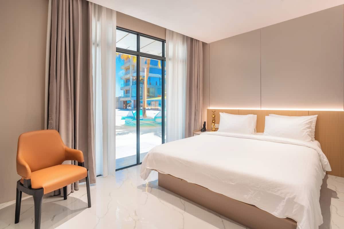 A modern hotel room with a large bed, a chair, and a view of a pool and buildings outside.