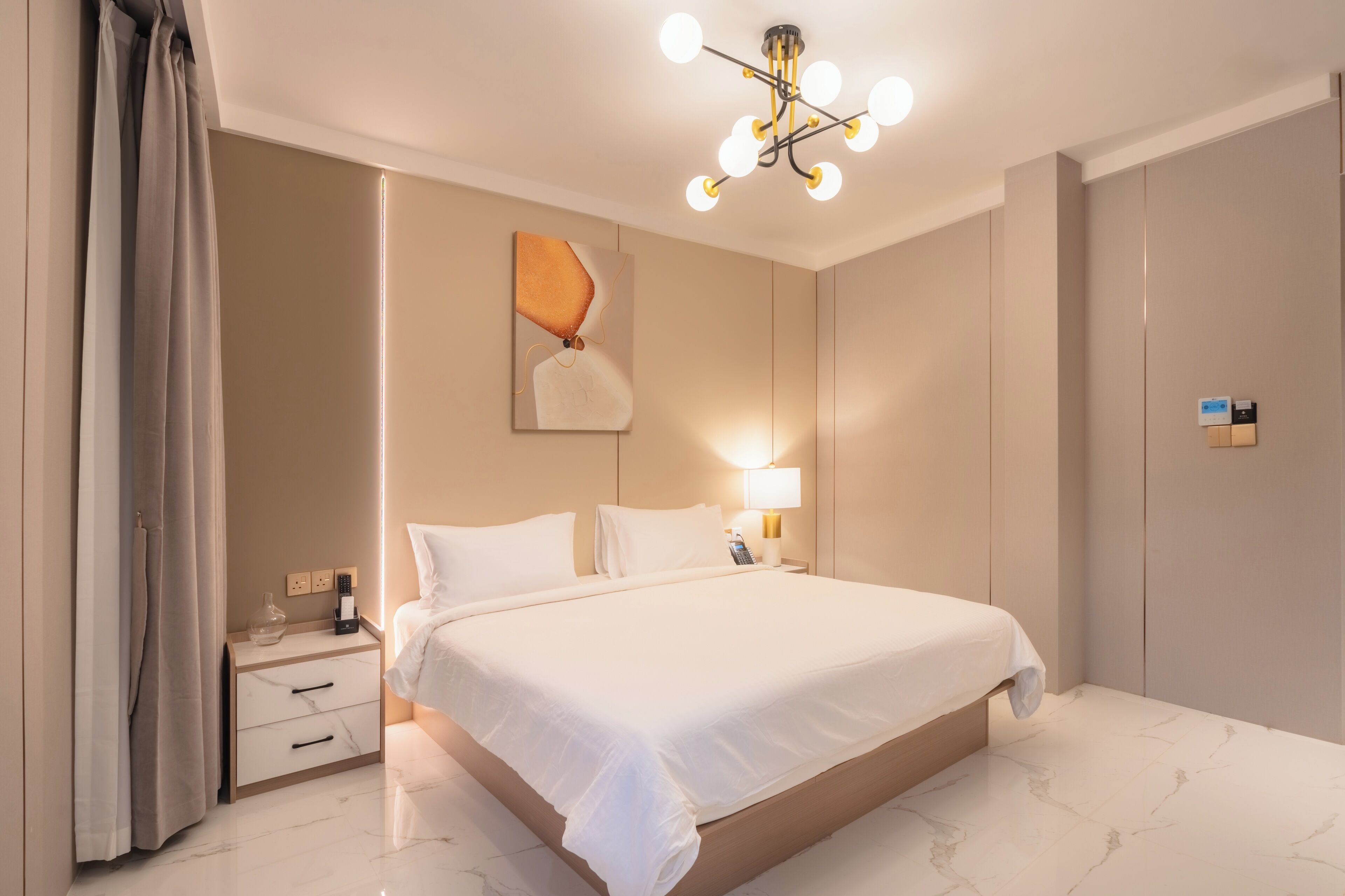 A modern hotel room with a large bed, bedside tables, a nightstand, a wall-mounted lamp, and a ceiling light fixture.