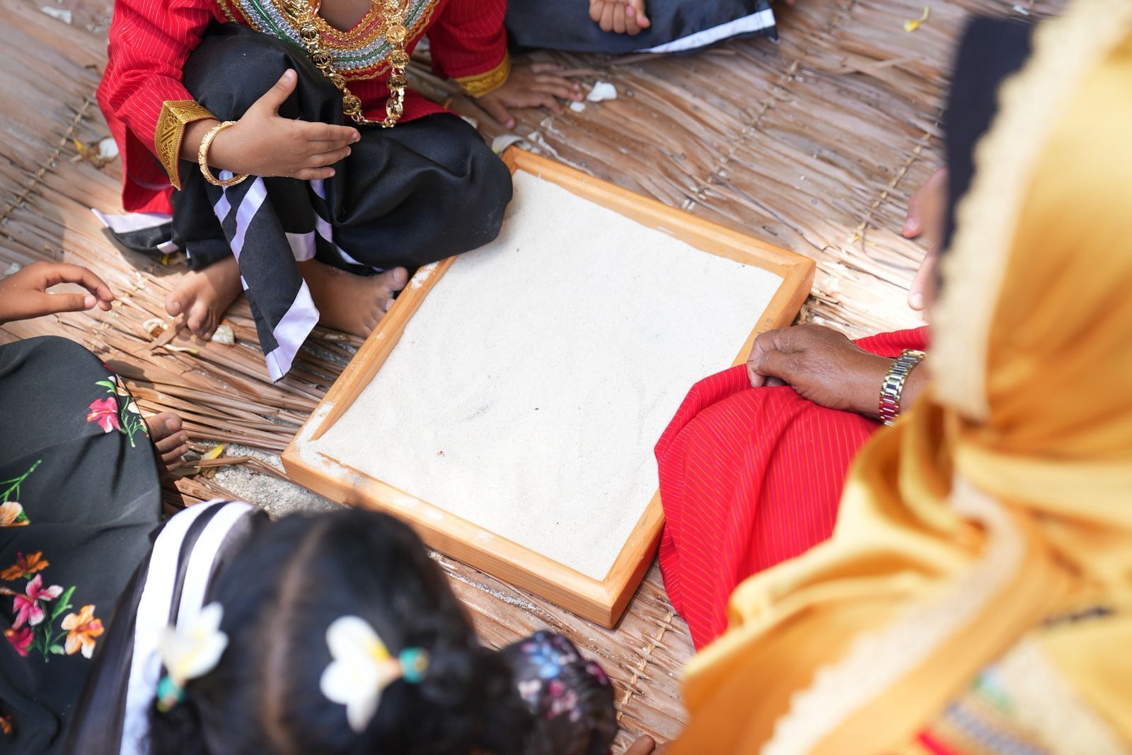 A group of people sitting on a woven mat, some wearing traditional attire.