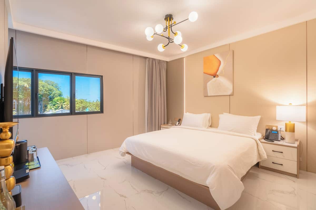 A hotel room with a large bed, a desk with a lamp, a telephone, and a view of trees through the window.
