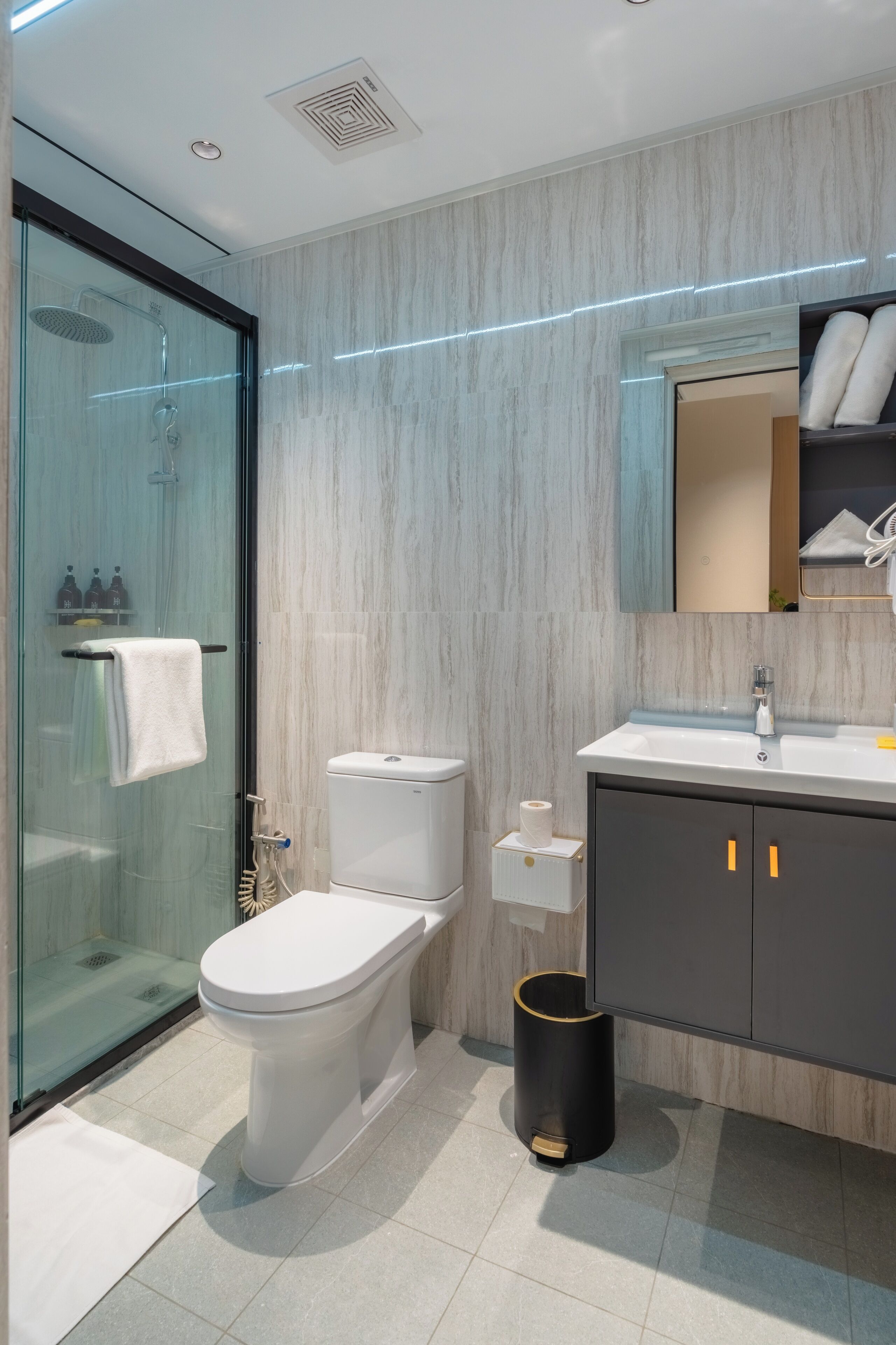 A modern bathroom with a glass shower enclosure, a toilet, a sink with a mirror above it, and a trash can.