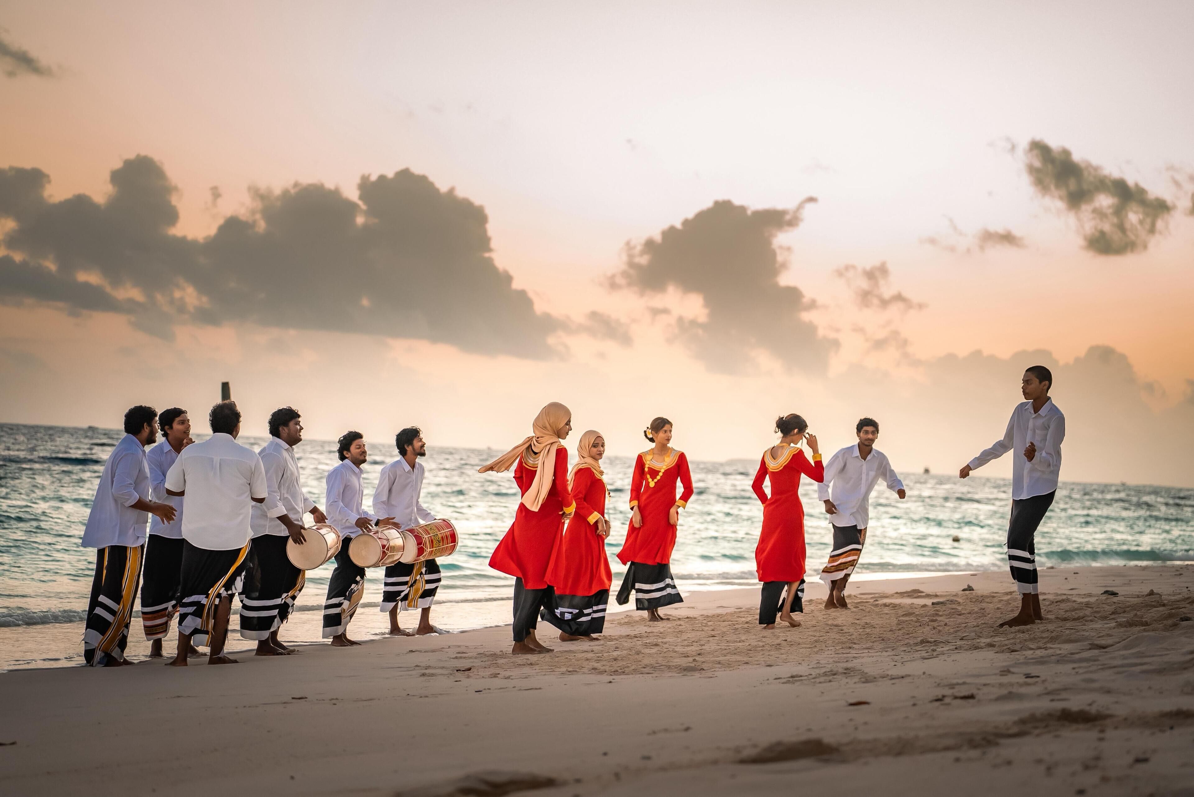 Een groep mensen op het strand, sommigen in witte overhemden en anderen in rode jurken, met een zonsondergang op de achtergrond.