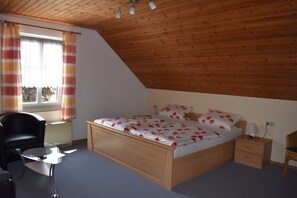 Ein Schlafzimmer mit Holzboden, einem Bett mit floralem Bettbezug, einem Nachttisch, einem Sessel und einem Fenster mit Vorhängen.