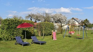 Ein grünes Areal im Freien mit einer Schaukel, einer Rutsche und zwei Liegestühlen unter einem roten Sonnenschirm.