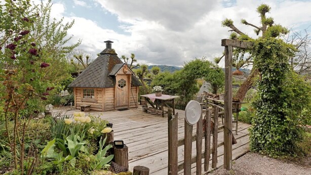 A rustic wooden cabin with a chimney, surrounded by lush greenery and a wooden deck.