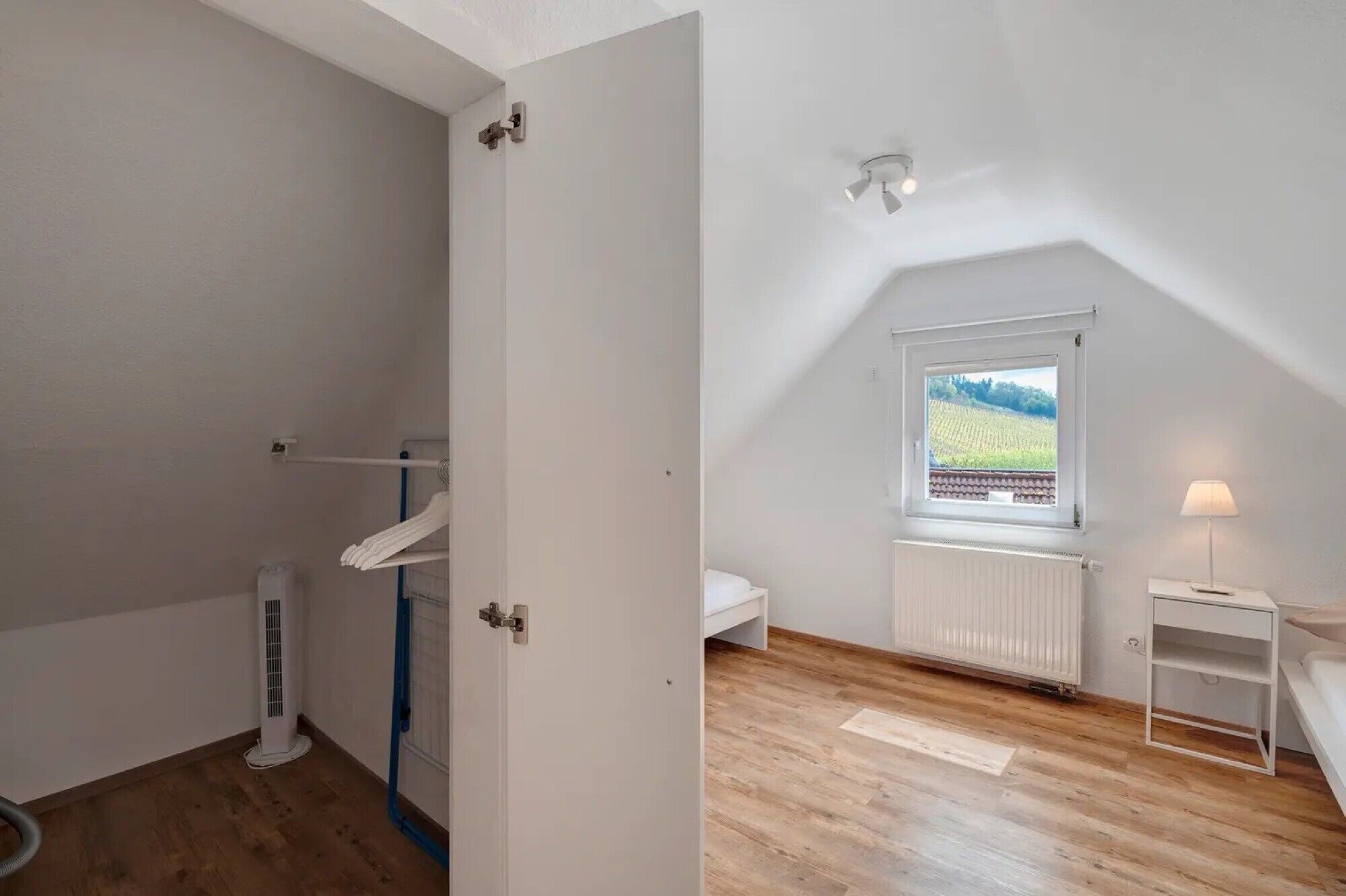 Ett rum med ett sluttande tak, ett fönster med utsikt över ett fält, ett element, ett litet bord med en lampa och en vit garderob.