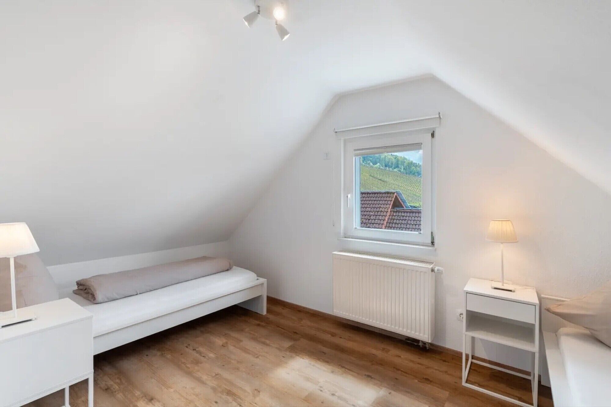 A small, white, sloped-roofed room with a bed, a radiator, a window with a view of greenery, and a small table with a lamp.
