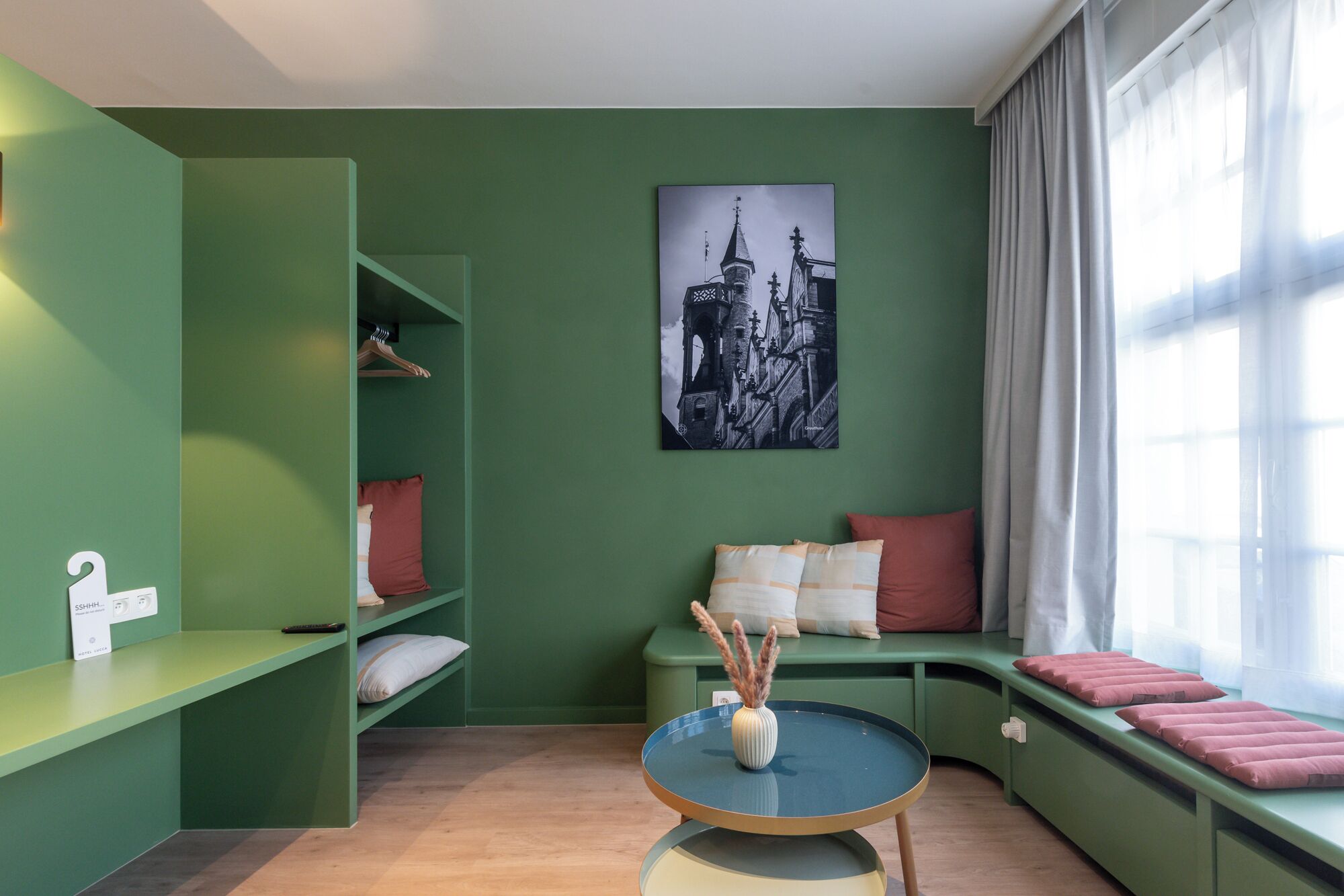 Una habitación con una pared verde, un cuadro de un paisaje urbano en blanco y negro, una mesa redonda con un jarrón y una ventana con cortinas.
