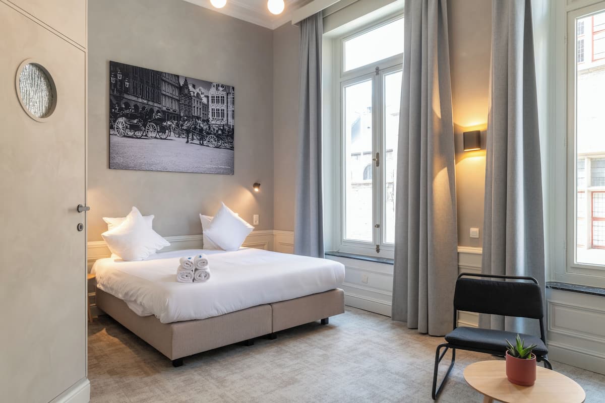 A bedroom with a bed, pillows, a chair, a small table with a plant, a wall-mounted light, and a painting.