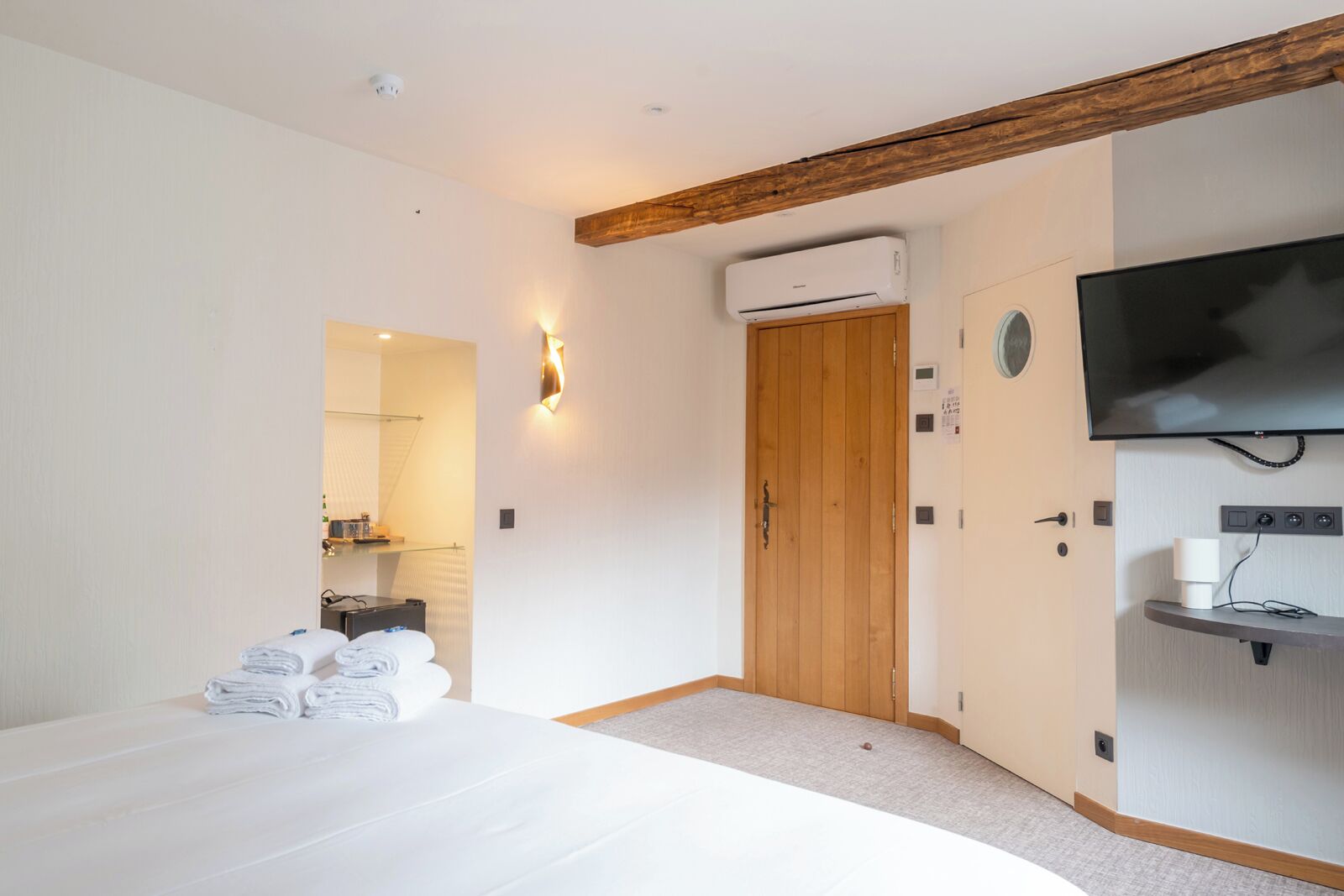 Habitación de hotel con cama, puerta de madera, unidad de aire acondicionado, televisor y una pequeña estantería con taza y vasos.