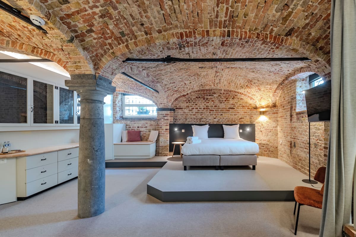 A bedroom with a brick ceiling, a bed, a nightstand, a chair, and a window.