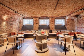 A brick-walled dining area with tables and chairs, a central serving station, and three windows. - Hotel Lucca (Bruges)